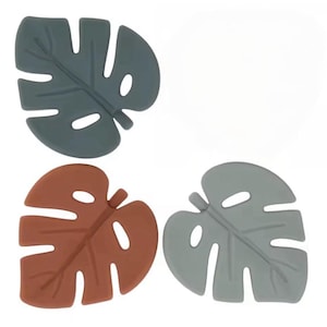 May include: Four leaf-shaped coasters in shades of gray and brown. The coasters are designed to resemble Monstera leaves, with cut-out holes and detailed veining. These decorative coasters are suitable for home decor.