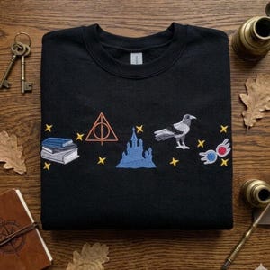 May include: Black sweatshirt with embroidered designs including books, a triangle, a castle, a bird, and glasses. The designs are in various colors, including orange, blue, and white, with yellow stars. The sweatshirt is folded on a wooden surface.