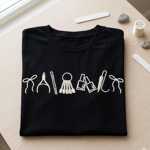 May include: Black t-shirt featuring white embroidered nail art tools. The design includes nail clippers, a cuticle pusher, nail polish bottles, and a nail file. The shirt is folded on a light-coloured wooden surface.