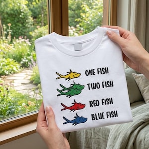May include: White t-shirt with embroidered fish in yellow, green, red, and blue. The text reads "ONE FISH, TWO FISH, RED FISH, BLUE FISH". The shirt is held up in front of a window with a green outdoor background.