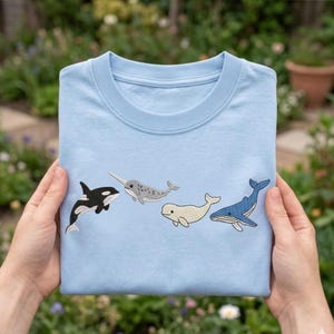 Puede incluir: Una camiseta azul claro con imágenes bordadas de una orca, un narval, una beluga y una ballena azul. Las ballenas son en negro, blanco, gris y azul. La camiseta está doblada y se sostiene frente a un fondo verde borroso.