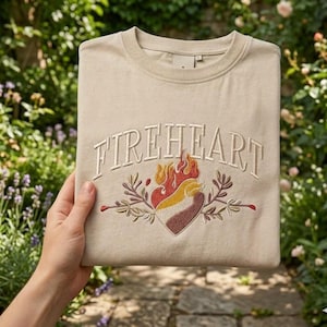 Könnte beinhalten: Beiges Sweatshirt mit dem gestickten Wort "FIREHEART" auf der Brust. Darunter befindet sich ein gesticktes Design eines Herzens mit Flammen und Blättern. Das Sweatshirt ist gefaltet und wird gehalten.