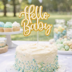 May include: A round, white frosted cake decorated with blue and white sprinkles and a gold glitter "Hello Baby" cake topper. The cake is on a white plate. Other party decorations are visible in the background.
