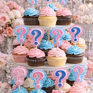 May include: A tiered dessert stand holds cupcakes with pink, blue, and yellow frosting. Each cupcake is topped with a glittery question mark, suggesting a gender reveal theme. The cupcakes sit on a silver stand, surrounded by flowers.