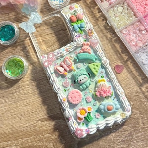 Puede incluir: Funda de teléfono transparente decorada con adornos en colores pastel. Presenta un conejito, una mariposa, flores y un letrero verde que dice "NICE DAY". La funda está rodeada por un borde blanco similar a glaseado.