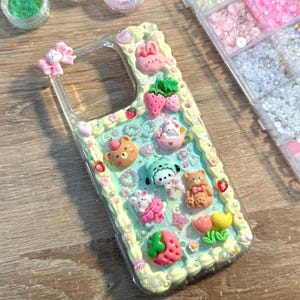 Puede incluir: Una funda transparente para teléfono decorada con varios abalorios 3D de colores. Los abalorios incluyen fresas, osos, conejos y otras figuras bonitas. La funda tiene un borde amarillo claro y está adornada con pequeñas perlas y gemas.