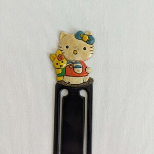 Może przedstawiać: Winietka Hello Kitty w stylu vintage. Zakładka ma czarny prostokątny kształt z wycięciem. Postać Hello Kitty ma na sobie czerwoną sukienkę z niebieską kokardą i towarzyszy jej żółty króliczy przyjaciel.