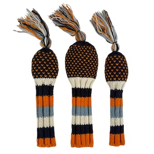 May include: Three golf club head covers. The covers are knitted with a navy blue base and orange accents. The top portion is navy blue with orange dots. The bottom has horizontal stripes of orange, light blue, and navy blue. Each cover has a tassel.
