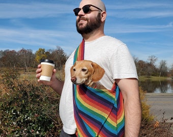 Può includere: Un uomo con occhiali da sole e una maglietta bianca porta un piccolo cane marrone e bianco in una tracolla arcobaleno. Tiene una tazza di caffè di carta marrone nella mano sinistra.