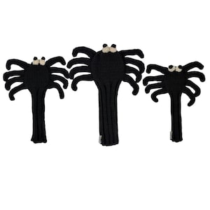 May include: Three black knitted spider-shaped golf club head covers. Each cover features a round body with eight extended legs and two small, cream-colored circular eyes positioned at the top. The covers have a long, ribbed neck section.