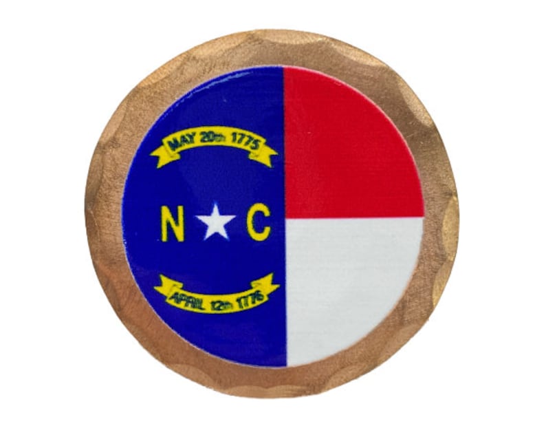 May include: A round, wooden coin with a gold-colored rim. The center features the North Carolina state flag, a blue field with a white star and the letters "N*C" in yellow. The flag is bordered by a red and white stripe. The words "MAY 20th 1775" and "APRIL 12th 1775" are written in yellow on a white ribbon.