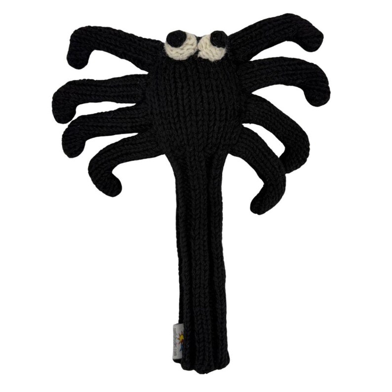 May include: A black knitted spider-shaped golf club headcover featuring eight long legs, a central body, and two round white eyes with black pupils. The item has a long, narrow neck section and a small white fabric tag attached to the lower edge.