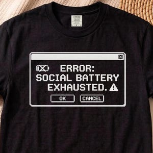 Op de afbeelding: Zwart T-shirt met een witte afbeelding die lijkt op een computerfoutmelding. De melding luidt "ERROR: SOCIAL BATTERY EXHAUSTED." met de knoppen "OK" en "CANCEL". Het shirt heeft een ronde hals en een klein label op de rug.