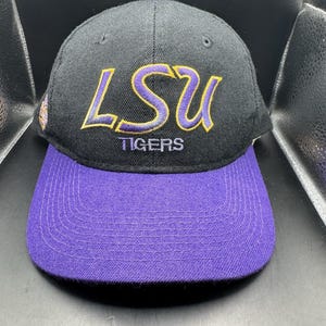 May include: Black baseball cap with a purple bill. The cap features the embroidered text "LSU TIGERS" in gold and purple. A classic, adjustable hat for fans.