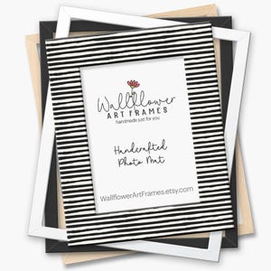 May include: A stack of picture frames in various colors, including black, white, and natural wood. The central frame features a black and white striped design and a white mat with the text "Wallflower Art Frames" and "Handcrafted Photo Mat."