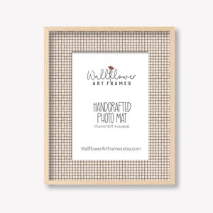 May include: A rectangular, light-colored wooden picture frame with a grid-patterned mat. The mat has a cream background with a black grid pattern. The frame contains the text "Wallflower Art Frames" and "Handcrafted Photo Mat".