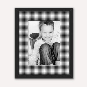 May include: A black rectangular picture frame featuring a white and black gingham patterned mat. The frame contains a black and white photograph of a smiling child wearing a long-sleeved shirt and jeans, with their knees pulled up.