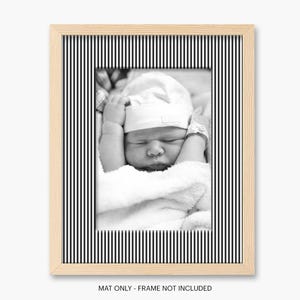 May include: A black and white photograph of a newborn baby in a white hat and blanket, framed in a light wood frame with a black and white striped border. The text at the bottom reads "MAT ONLY - FRAME NOT INCLUDED."