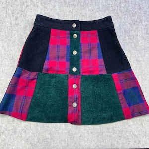 May include: A-line skirt featuring a patchwork design with a black waistband. The skirt showcases a vibrant mix of red, green, and blue plaid patterns, accented by a central panel of green fabric. Silver buttons run down the front.