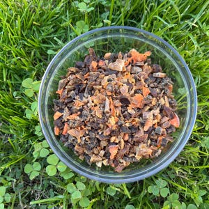 May include: A clear glass bowl containing a mixture of dried, chopped ingredients, with orange and brown hues. The bowl is placed on a bed of green grass with clover.