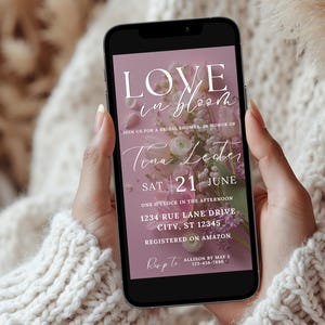 May include: A smartphone displays a bridal shower invitation with the text "LOVE in bloom." The invitation features floral arrangements in pink and white, with details including the date, time, and location. The phone is held against a soft, textured background.