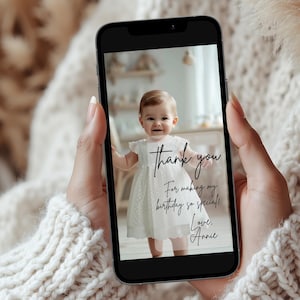 May include: A smartphone displays a photo of a baby in a white dress. The photo includes the text "thank you" and "For making my birthday so special! Love, Annie."