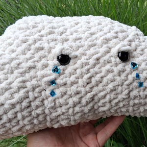 May include: A plush cloud-shaped pillow crafted from off-white, textured yarn. The pillow features two black, round eyes with three small, blue, teardrop-shaped rhinestones cascading from each eye, mimicking the appearance of falling tears.