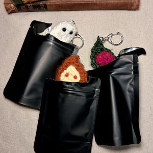 May include: Three small crocheted figures attached to silver-toned keyrings, each partially tucked inside a black, rectangular, resealable foil pouch. The figures include a white ghost, a brown-haired character, and a dark green leaf-shaped item.