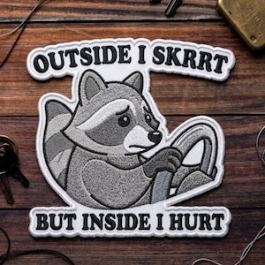 May include: Embroidered patch featuring a raccoon driving a car, with the text "OUTSIDE I SKRRRT BUT INSIDE I HURT" in black letters on a white background. The raccoon is gray and white, with a sad expression.