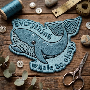 May include: Embroidered patch featuring a blue whale with the text "Everything whale be okay." The patch is surrounded by a blue border. Other sewing supplies, including thread, buttons, and scissors, are visible.