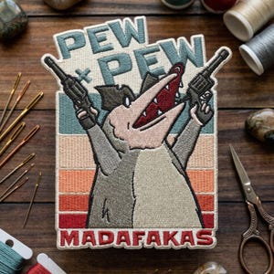 May include: Embroidered patch featuring a cartoon opossum holding two guns. The patch has the words "PEW PEW MADAF*KAS" in a retro color scheme of beige, blue, orange, and red. The opossum is gray and pink.