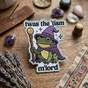May include: Embroidered patch featuring a green frog dressed as a wizard, wearing a purple hat and cape, holding a staff. The patch has the text "twas the 'tism" and "m'lord". The patch is on a wooden surface.