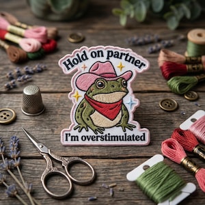 May include: Embroidered patch featuring a cartoon frog wearing a pink cowboy hat and red bandana. Text on the patch reads "Hold on partner, I'm overstimulated." The patch is surrounded by embroidery floss, scissors, and buttons.