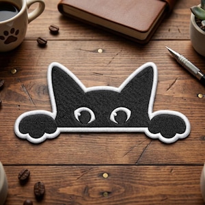 May include: Black cat patch with white embroidered outline. The cat's head and paws are visible, with large, open eyes. The patch is on a wooden surface, with coffee beans and a mug in the background.