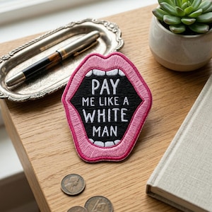May include: Embroidered patch with a pink outline of open lips, black interior, and white teeth. The text "PAY ME LIKE A WHITE MAN" is in white. The patch is on a wooden surface, with a pen, tray, succulent, and book nearby.