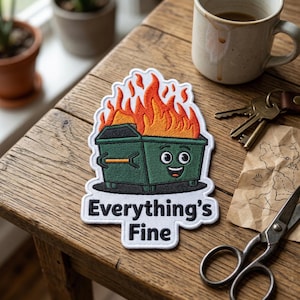 May include: Embroidered patch featuring a green dumpster on fire with orange flames. The dumpster has a smiling face and the text "Everything's Fine" in white. The patch is on a wooden surface.