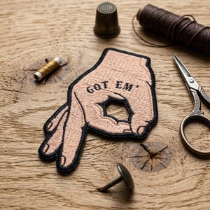May include: Embroidered patch in the shape of a hand making the 'okay' gesture, with the words "GOT EM" stitched in black. The patch is peach-colored with a black border. Sewing tools, including scissors and thread, are arranged around the patch on a wooden surface.