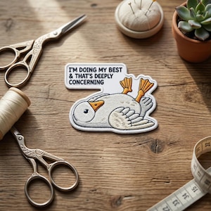 May include: Embroidered patch featuring a cartoon goose lying on its back with the text "I'M DOING MY BEST & THAT'S DEEPLY CONCERNING." The patch is white, gray, and orange. Sewing supplies, including scissors and thread, are in the background.