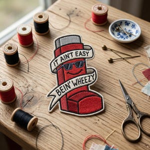 May include: A red and black embroidered patch with the text "IT AIN'T EASY BEIN' WHEEZY" and a cartoon inhaler wearing sunglasses. The patch is surrounded by spools of red and black thread, a small dish with pins, and a pair of scissors.
