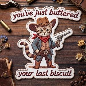May include: Embroidered patch featuring a cat dressed as a cowboy, holding a gun. The patch has the text "you've just buttered your last biscuit" in a western-style font. The cat wears a brown hat, red bandana, and cowboy boots.