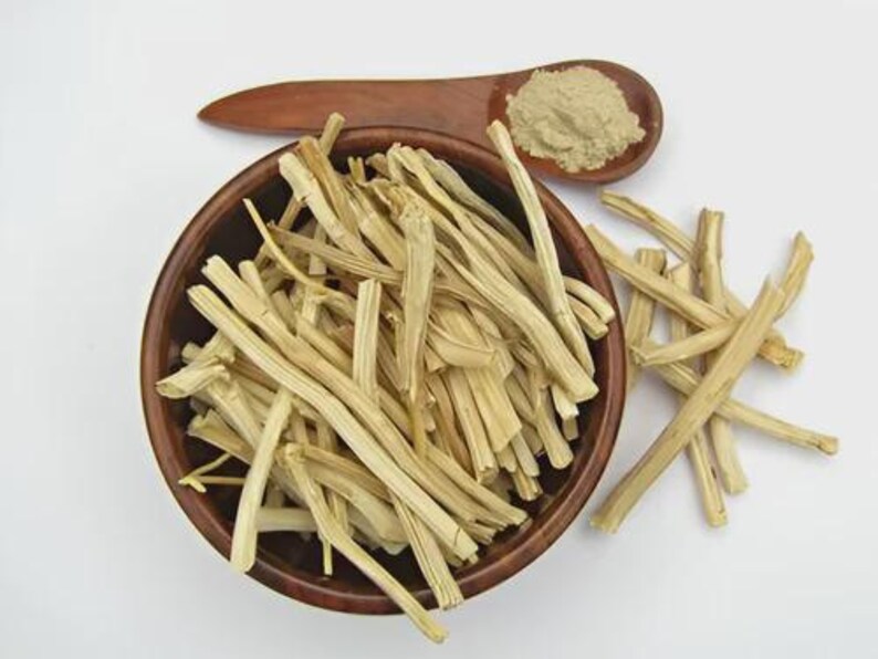 May include: A wooden bowl filled with light-colored, dried plant stems. A wooden spoon holds a pile of light brown powder. The items are on a white surface. The bowl and spoon are made of dark brown wood.