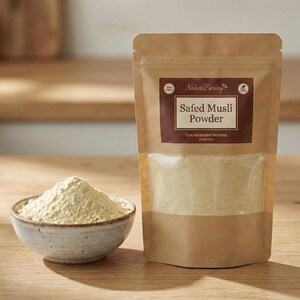 May include: A package of Safed Musli Powder from NatureSpring, with a bowl of powder in front. The package is brown with a dark brown label. The bowl is small and light-colored, filled with a light yellow powder. The background is a wooden surface.