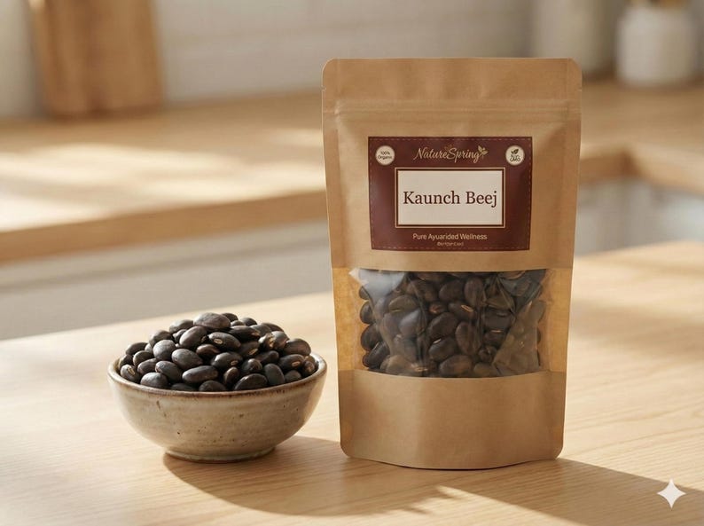 May include: A package of Kaunch Beej seeds with a clear window, next to a small bowl filled with similar seeds. The package has a brown label with the text "Nature Spring" and "Kaunch Beej". The seeds are dark brown.