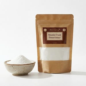 Puede incluir: Una bolsa de papel marrón de edulcorante de fruta del monje con una ventana transparente y un pequeño cuenco de cerámica lleno de polvo blanco. La bolsa tiene una etiqueta marrón con el texto "Monk Fruit Sweetener". El fondo es blanco.