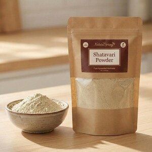 May include: A package of Shatavari Powder from Nature Spring, with a clear window showing the powder inside. A small bowl of the powder sits next to the package. The text on the package reads "Shatavari Powder, Pure Ayurvedic Wellness."