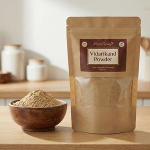 May include: A brown paper bag of Vidarikand Powder with a clear window, and a wooden bowl filled with the same powder. The bag has a brown label with white text. The background includes wooden kitchenware and white jars.