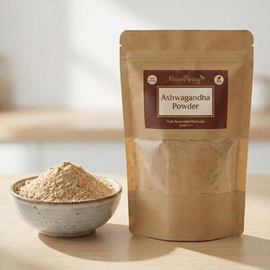 May include: A brown paper bag of Ashwagandha Powder with a clear window, and a small ceramic bowl filled with the powder. The bag has a brown label with the text "Ashwagandha Powder" and "Pure Ayurvdeda Wellness". The background is a light-colored wooden surface.