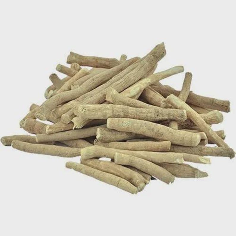 Op de afbeelding: Een stapel lichtbruine, gedroogde ashwagandha-wortels. De wortels vari&euml;ren in grootte en vorm, met een ruwe, getextureerde uitstraling. De afbeelding is genomen tegen een effen, lichte achtergrond, die de natuurlijke kleur en textuur van de wortels benadrukt.