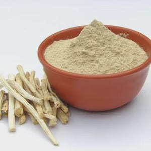 May include: A terracotta bowl filled with a light brown powder sits next to a pile of light-colored, dried sticks. The powder is finely ground, and the sticks have a rough texture. The items are set against a plain white background.