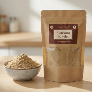 May include: A brown paper bag and a small ceramic bowl filled with Akarakara powder. The bag has a clear window and a label that reads "Akarakara Powder" and "Pure Ayurvedic Wellness". The bowl is light brown and sits on a wooden surface.